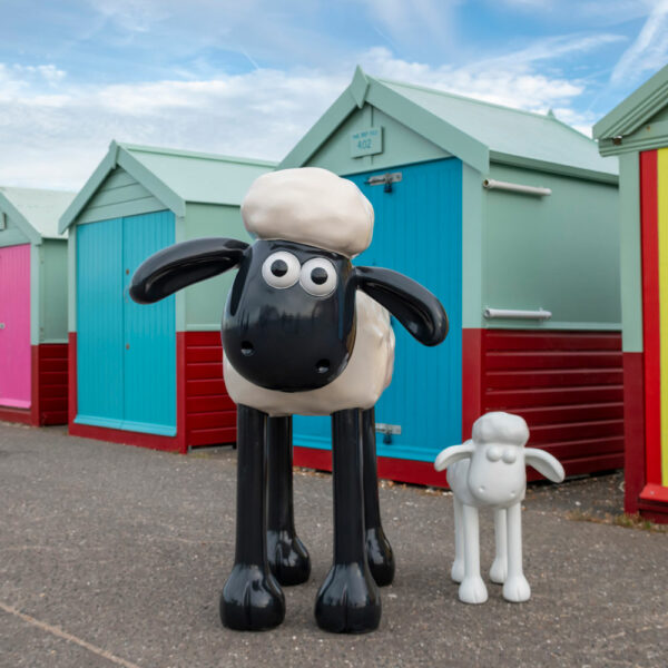 A big shaun the sheep sculpture with a smaller blank shaun the sheep sculpture outside of some beach huts